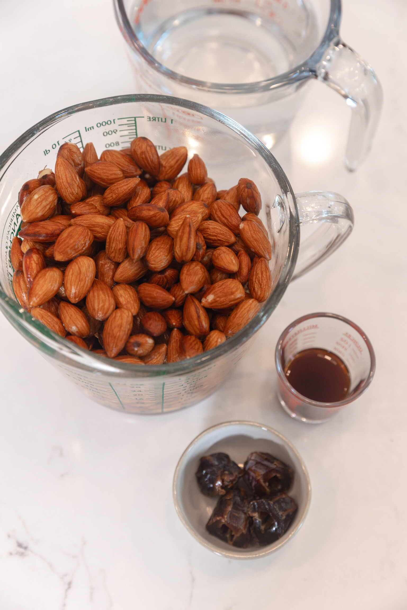 Measuring cup of raw almonds next to measuring cup of water, with a small cup of dates and small cup of vanilla extract.