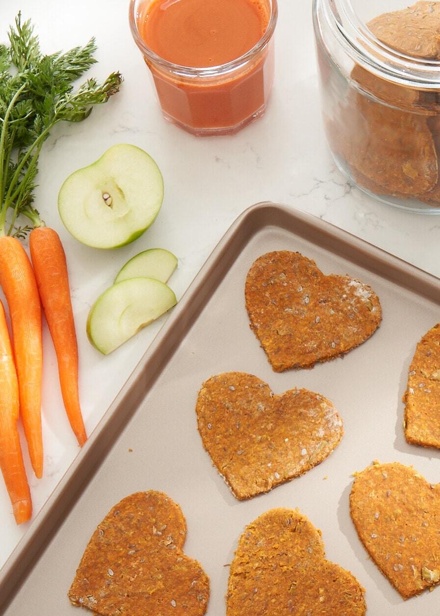 heart shaped dog treats on a parchment-paper-covered cookie sheet, next to carrots and sliced green apples.