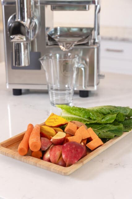 A wooden tray holds ingredients for sweet potato juice: apples, carrots, sweet potatoes, yellow beets, and romaine, in front of a PURE Juicer.