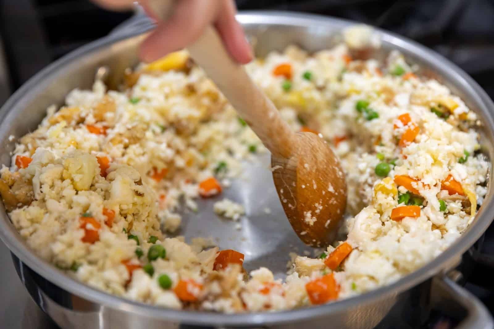 A wooden spoon stirs vegan cauliflower fried rice made with homemade cauliflower rice, carrots, peas, ginger, garlic, and egg substitute.