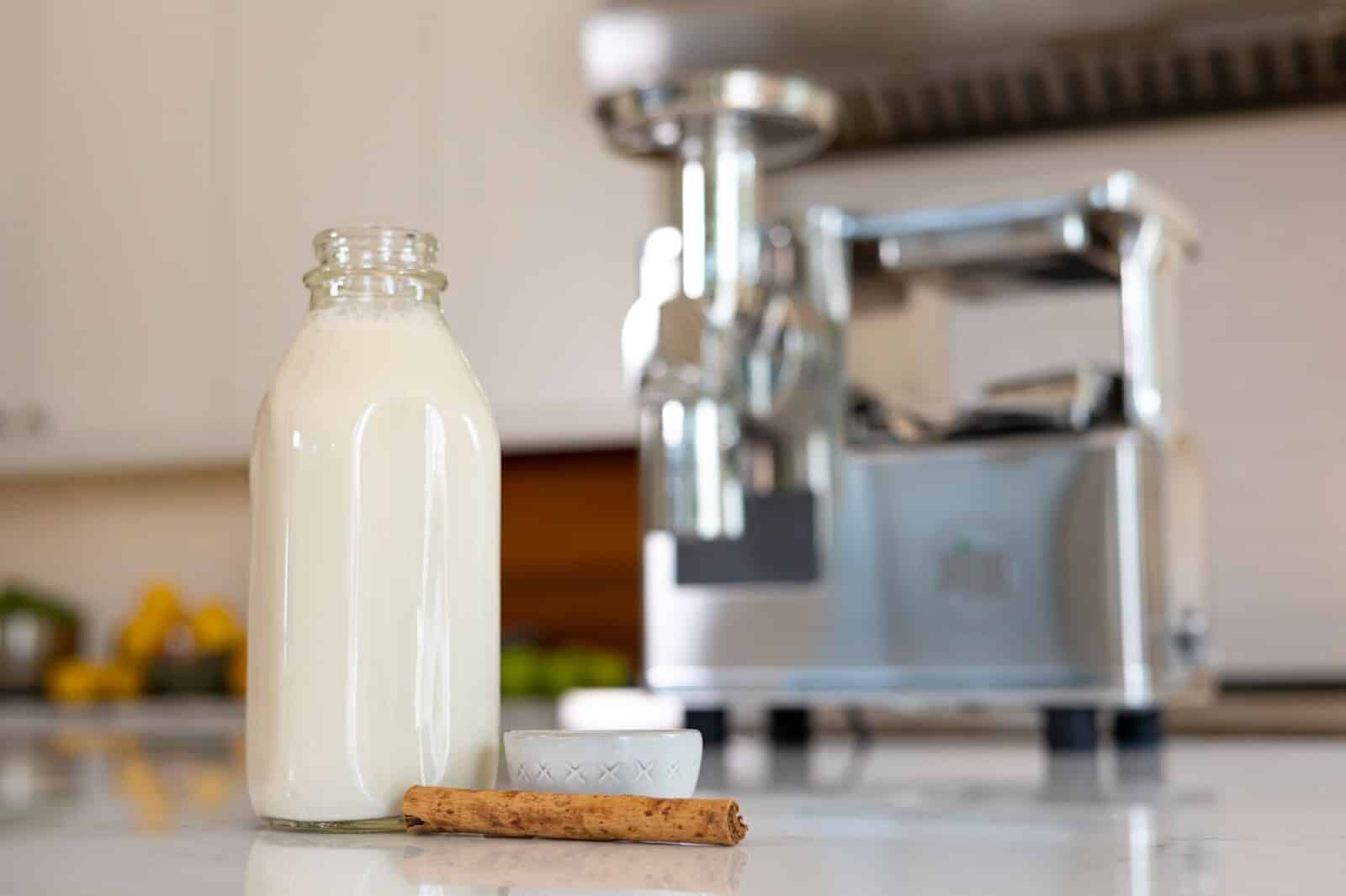 A glass jar of tiger nut milk sits beside a cinnamon stick and small white bowl; the PURE Juicer stands in the background.