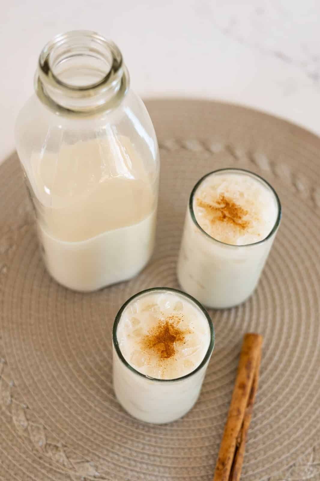 Two glasses of creamy tiger nut milk, garnished with ice cubes and sprinkles of cinnamon, rest on a placemat between a large glass jar of tigernut milk and a cinnamon stick.