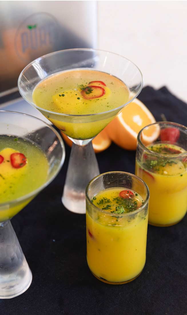 Two glasses of orange-chili-matcha juice sit on a black napkin, in front of two chilled martini glasses holding servings of orange-chili-mocktails.