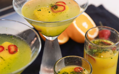 Two glasses of orange-chili-matcha juice sit on a black napkin, in front of two chilled martini glasses holding servings of orange-chili-mocktails.