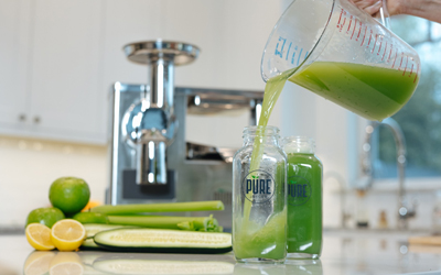 Refreshing cucumber celery juice sweetened with green apples and the subtle tang of lemon juice is poured from a pitcher into a glass bottle labeled PURE Juicer. Two juice bottles stand in front of a PURE Juicer, sliced lemons, sliced cucumbers, and a stack of green apples.