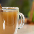 A glass mug full of warm, mulled apple cider, garnished with a cinnamon stick, sits on a countertop